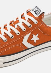 Oranssi kangastossu, jossa on valkoiset metalliset reikäkappaleet, valkoiset nauhat, valkoinen tähtilogo ja raidoitettu kumipohja mustilla yksityiskohdilla.