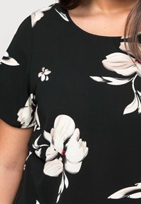 Czarna bluzka z krótkim rękawem z dużymi białymi wzorami kwiatowymi i subtelnymi czerwonymi akcentami na górnej części ciała i ramieniu osoby.