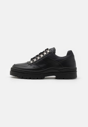Chaussure basse en cuir noir avec semelle épaisse à crampons, œillets métalliques et lacets noirs, vue de profil sur fond blanc.