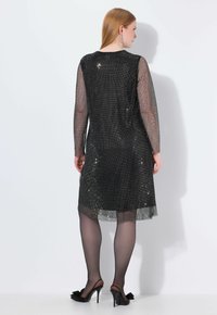 Schwarzes Pailletten-Kleid mit langen transparenten Ärmeln, rundem Ausschnitt und einer Länge unterhalb des Knies. Betont durch schimmernde Pailletten auf der gesamten Fläche.