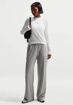 Kvinde stående iført hvid Nike langærmet trøje, grå vidde-bukser, hvide sneakers, sort skuldertaske og hvidt pandebånd.