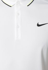 Biała koszulka polo z teksturowanej tkaniny, z zapięciem na dwa guziki, czarnym logo Nike i kołnierzykiem z czarnym i neonowo-żółtym akcentem.