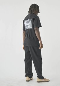 Mann mit Dreadlocks, der ein schwarzes Grafik-T-Shirt, lockere schwarze Hosen und beige Sneakers trägt und mit dem Rücken zur Kamera steht, während er über die Schulter auf einem weißen Hintergrund schaut.