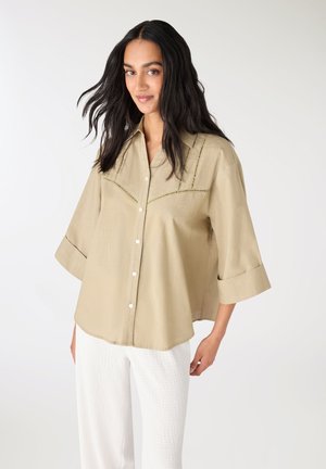 Donna con lunghi capelli neri indossa una camicia beige ampia con bottoni e maniche arrotolate e pantaloni bianchi testurizzati, in piedi davanti a uno sfondo semplice.