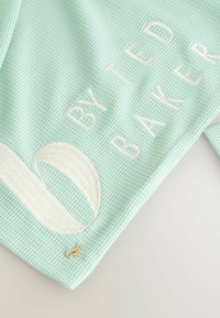 Světle mátově zelená texturovaná látka s vyšitým bílým textem "BY TED BAKER" a detailním kruhovým logem, doplněná malým zlatým akcentem.