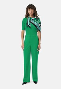 Haut en maille côtelée vert et pantalon large, tous deux dotés d'une texture lisse. Une écharpe à motifs tombe sur l'épaule. Talons noirs pointus.