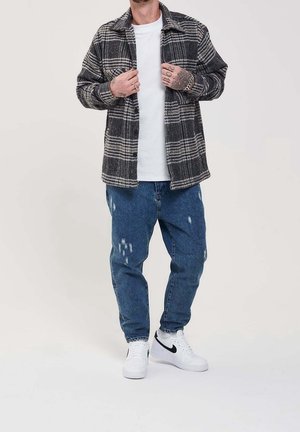 Homme portant une veste à carreaux noire et blanche par-dessus une chemise blanche, jean bleu déchiré, baskets blanches Nike, debout en ajustant sa veste avec les mains.