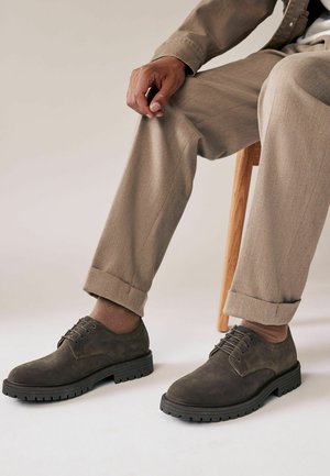Dunkelgraue Wildlederschuhe mit runder Spitze, Schnürdesign und dicker schwarzer Gummisohle, kombiniert mit hellbeigen Hosen.