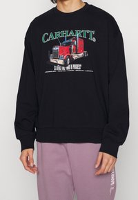 Sudadera negra con un gráfico de un camión rojo, logotipo verde y texto. Puños y dobladillo acanalados, corte relajado, fabricada en tejido suave.