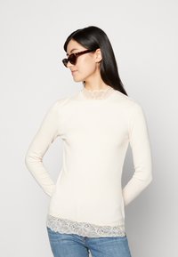 Langermet kremfarget ribbekjorte med blondekant rundt hals og nederkant; kombinert med jeans og mørke solbriller. Glatt tekstur, tettsittende design.