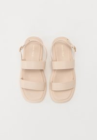 Beige flat sandals featuring two wide straps and a thin adjustable ankle strap with a gold-tone buckle. Smooth, minimalistic design.