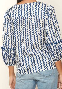 Bílý top s modrým cikcak vzorem, tříčtvrteční rukávy s vyšívanými detaily a bambulkami na manžetách. Měkká, lehká tkanina.