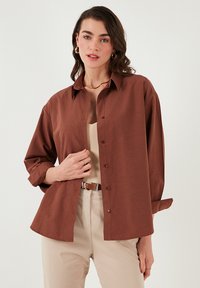 Brown button-up shirt with a relaxed fit, long sleeves rolled to the elbows, and a soft cotton-like texture. Features a classic collar and subtle stitching.