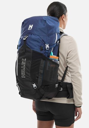 Personne portant une veste beige et un short noir transporte un grand sac à dos de randonnée Millet bleu et noir avec une bouteille d'eau dans la poche latérale.