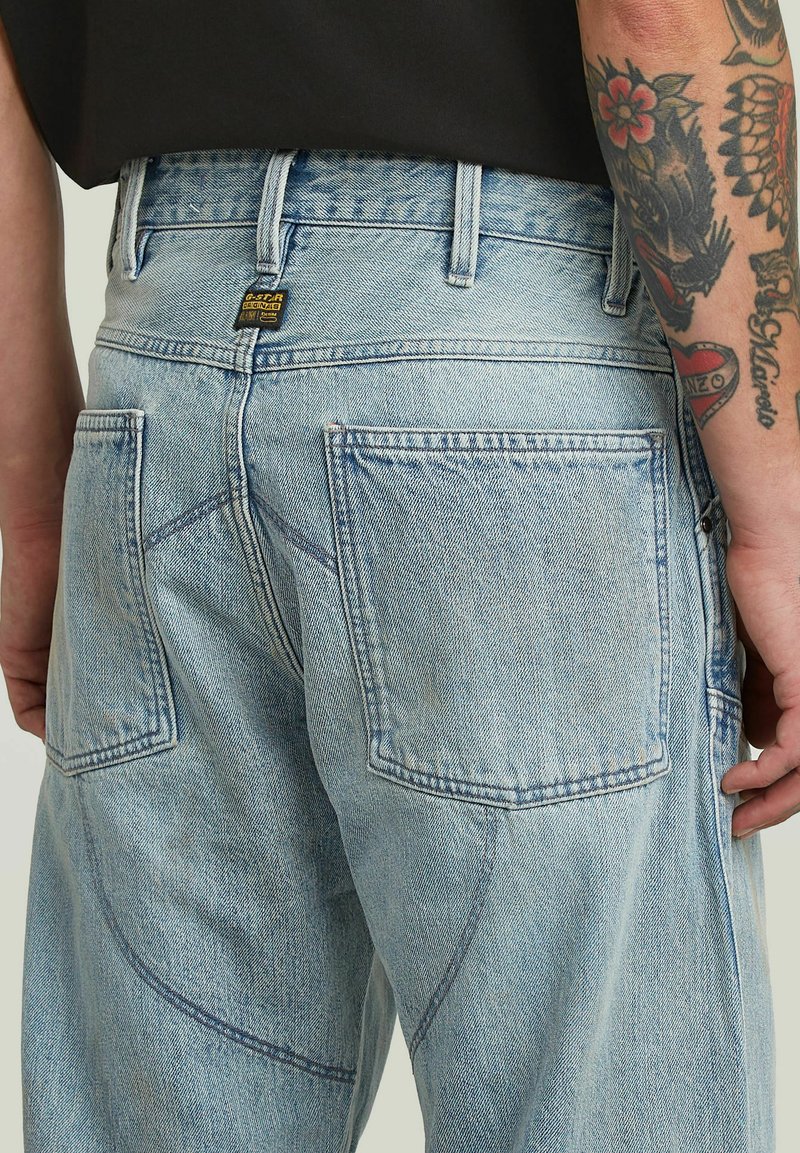 Jeans in denim celeste con grandi tasche posteriori e dettagli delle cuciture ben visibili, indossati da una persona con un braccio tatuato e una maglietta nera.