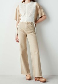 Persona che indossa jeans larghi beige, una maglietta beige a maniche corte, un gilet di maglia bianco con frange e zoccoli di tela color tortora con borchie.