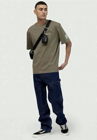 Olivenfarvet t-shirt med tekst, parret med mørkeblå løse jeans. En sort crossbody taske bæres, og sorte sneakers har hvide accenter.