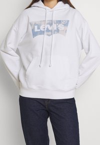 Bílá mikina s kapucí a předními klokankovými kapsami, se grafickým logem Levi's v modro-bílé barvě. Materiál z bavlny, uvolněný střih.