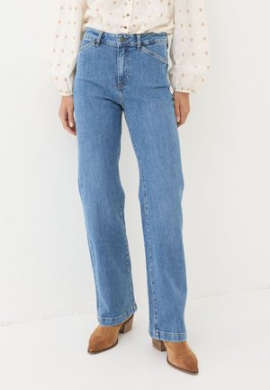 Person wearing light blue straight-leg jeans, beige patterned blouse, and tan suede heeled ankle boots standing against a white wall.