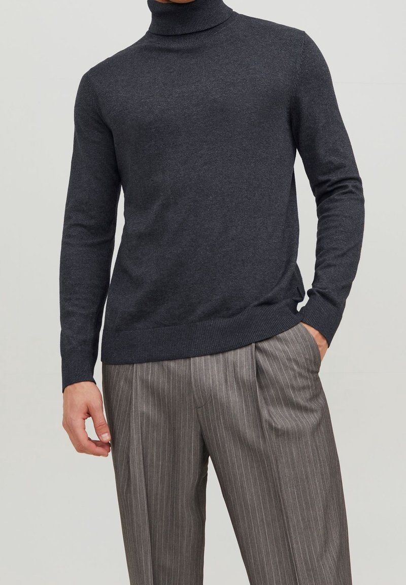 Homme portant un pull à col roulé gris foncé et un pantalon gris à fines rayures, une main dans la poche et l'autre détendue sur le côté.