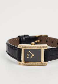 Relógio retangular dourado com mostrador preto, detalhes dourados e acabamento em cristal. Pulseira de couro preto com textura de crocodilo e fechos dourados.