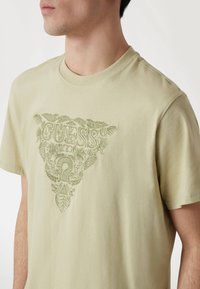 Šviesiai žalia trumpomis rankovėmis marškinėliai su tekstūriniu grafiniu spaudiniu, kuriame yra "GUESS" logotipas ir gėlių raštai, apvaliu kaklu ir minkšta medžiaga.