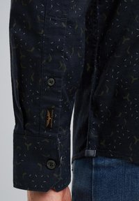 Chemise à motifs marins avec un poignet boutonné et une étiquette, des motifs géométriques discrets en vert et blanc, et un tissu texturé.