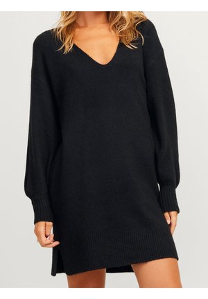 Femme portant une longue robe-pull noire à col en V avec des poignets et un ourlet côtelés, debout devant un fond clair uni.
