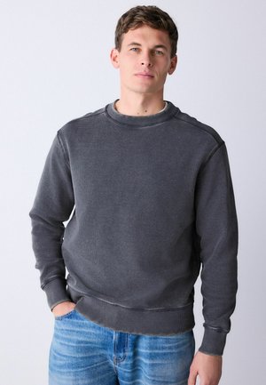 Gray sweatshirt in soft, textured fabric, featuring a ribbed crew neck and cuffs, with a relaxed fit and minimal seams. Worn with blue jeans.