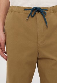 Pantalon marron avec taille élastique et cordon bleu noué en un nœud, porté avec une chemise blanche rentrée à l'intérieur.