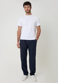 Hombre con cabello rizado, vestido con camiseta blanca, pantalones azul marino y zapatillas blancas, de pie frente a un fondo gris claro.