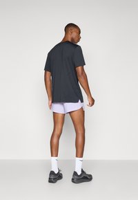 Schwarzes kurzärmliges T-Shirt für Herren mit Rundhalsausschnitt und kleinem Schlitzdetails auf der Rückseite, kombiniert mit helllila Shorts und schwarzen Sportschuhen.
