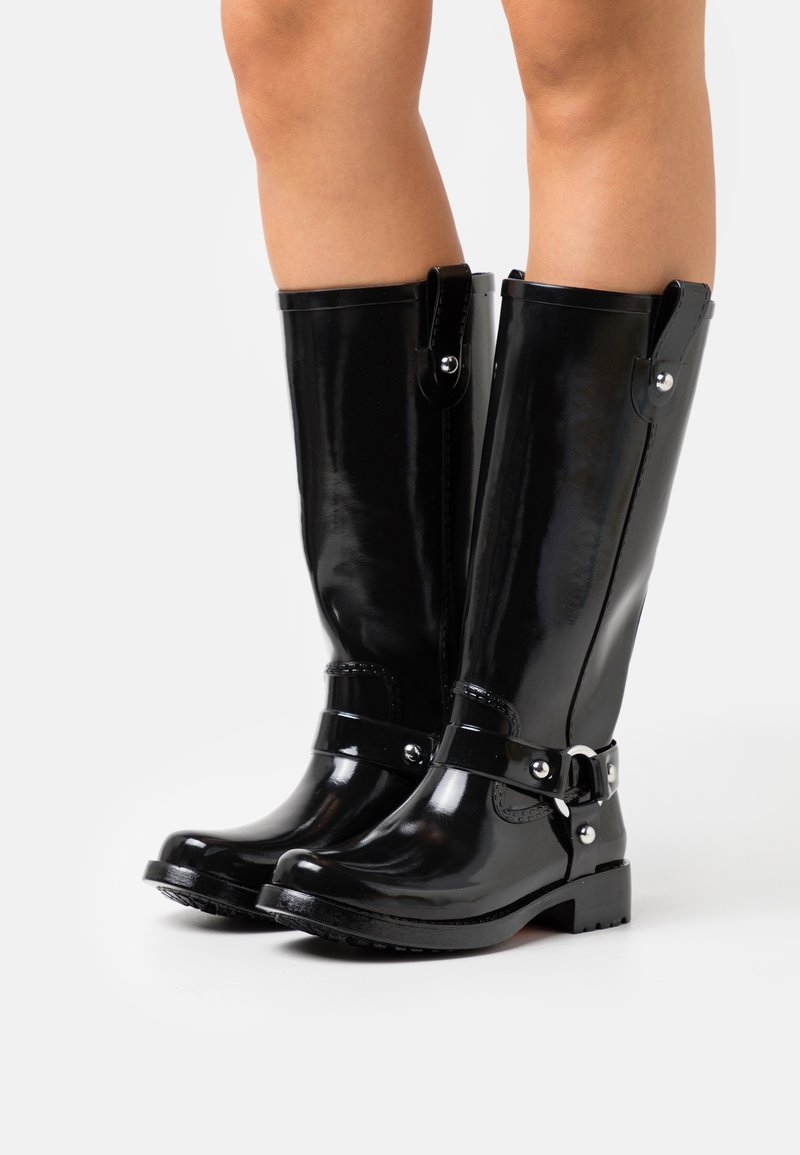 Bottes noires brillantes montant jusqu'au genou avec des sangles et des boucles argentées portées sur des jambes nues sur un fond blanc uni.