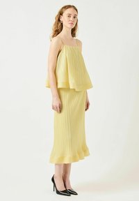 Vestido amarillo plisado con diseño en capas, tirantes finos y dobladillo acampanado. Combinado con zapatos de tacón alto negros. Silueta simple y elegante.