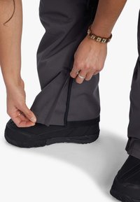 Grå utomhusbyxor med en zippered ankelsöppning, justeras av en hand. Svarta vattentäta stövlar med texturerade sulor är synliga.