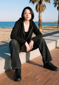Fiatal nő fekete kapucnis pulcsiban és farmerben, aki egy betonperemen ül a tengerpartnál, háttérben pálmafák és az óceán.