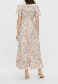 Femme portant une robe longue à fleurs, à manches courtes avec un ourlet volanté et des sandales à talons beiges, se tenant devant un fond uni.