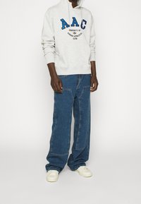 Grå huvtröja med "AAC"-logotyp i blått, tillsammans med löst sittande blå jeans och vita sneakers. Materialet verkar mjukt och har en avslappnad passform.