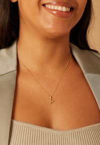 Collier en or avec un petit pendentif en forme de lettre « Y », porté sur un cou dénudé au-dessus d'un vêtement texturé de couleur claire.