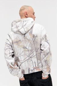 Sweatshirt com capuz em branco com um estampado texturizado inspirado na natureza, apresentando ramos e folhas em tons de bege e cinza.