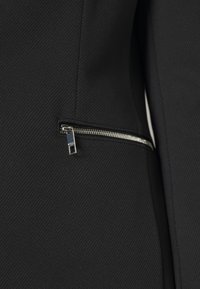 Sezione di giacca sartoriale nera con tessuto strutturato, dotata di una tasca con cerniera metallica sul lato sinistro. La cerniera è di colore argento.