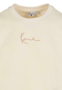 Cream cotton T-shirt featuring a round neckline and subtle embroidered logo in a tan color on the front. Soft texture with casual fit.