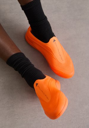 Oransje slip-on sko med teksturert strikket overdel, som ligger over svarte ribbede sokker på en betongoverflate. Har merkevarelogo i svart.