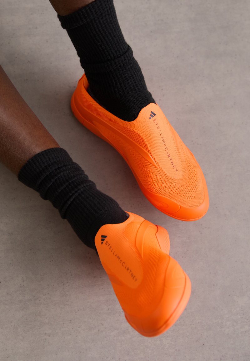 Pantofi portocalii fără sireturi, cu materiale tricotate texturate, așezați deasupra șosetelor negre cu riglaje pe o suprafață de beton. Prezintă logo-ul mărcii în negru.