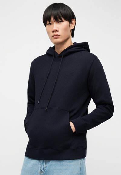 Νεαρός φοράει σκουρόχρωμο μπλε φούτερ με κουκούλα και έχει τα χέρια στην μπροστινή τσέπη, φοράει ανοιχτόχρωμο μπλε τζιν, στέκεται μπροστά σε ένα μονόχρωμο φόντο.