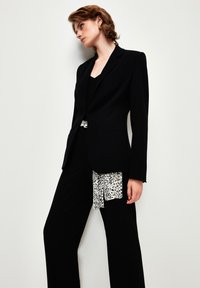 Blazer nero su misura con rever a punta, abbinato a pantaloni neri morbidi accentuati da una sciarpa con stampa animalier bianca e nera. Texture liscia.