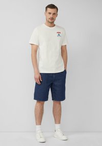 T-shirt en coton blanc avec un design graphique bleu sur la poitrine, associé à un short en denim bleu foncé et des baskets blanches. Style simple et décontracté.