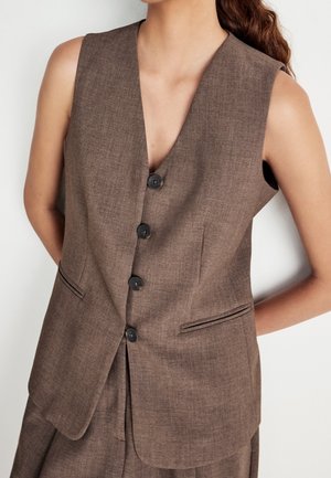 Brązowa, bezrękawnikowa kamizelka na guziki z dwoma przednimi kieszeniami bocznymi, noszona przez osobę na jednolitym tle.