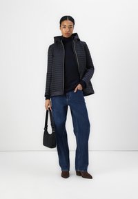 Veste noire matelassée avec capuche, fermeture éclair à l'avant et poignets côtelés, associée à un pull sombre, un jean bleu à jambes larges et des bottes à talons marron.