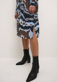 Pitkähihainen mekko, jossa on ruskea, sininen ja musta abstrakti kuvio, sivuaukko, yhdistetty mustiin nilkkureihin, joissa on paksu korko.